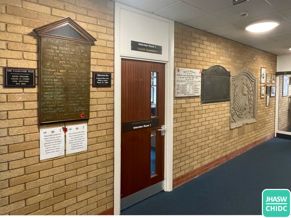 A photograph showing a carpeted corridor in the council offices, with about eight plaques of different sizes on the wall either side of a meeting room door, and some framed photo-graphs further on.